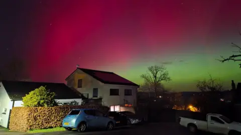Scarlet, pink and green aurora above houses and cars in Dartington, Devon