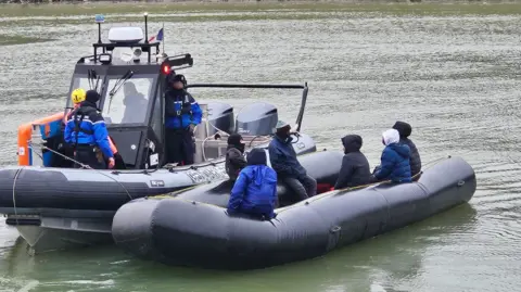 French police alongside a grey inflatable 'taxi-boat' on the Aa canal in Gravelines; men seated on the dinghy as it is towed toward the dock.